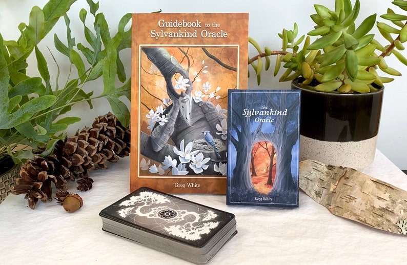 May include: A set of oracle cards with a guidebook and a smaller book titled "The Sylvankind Oracle". The guidebook has a brown cover with a colorful illustration of a tree spirit with white flowers. The smaller book has a blue cover with a forest scene.