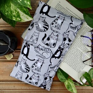 May include: A rectangular fabric pouch featuring a repeating pattern of black and white cats on a light gray background. The pouch has a folded front pocket design and is constructed from a soft textile material with a consistent cat print throughout.