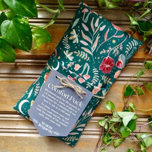 May include: A dark green comfort pack with a floral pattern in red, pink, and white. The pack is tied with twine and has a tag that reads "Comfort Pack" and "Warm Regards". The background includes green plants.
