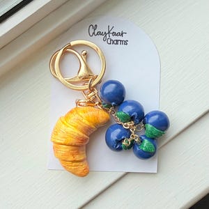 Peut inclure: Un porte-clés doré avec une breloque croissant doré-brun et un groupe de quatre breloques myrtilles bleues brillantes avec de petites feuilles vertes, attaché à un anneau doré. La carte présente le texte ClayKaar Charms en noir.