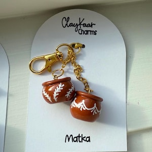 May include: A keychain featuring two miniature brown pots with white floral designs. The pots are connected by a gold chain and attached to a gold clasp. The card reads "ClayKaar Charms" and "Matka".