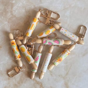 May include: A collection of miniature rolling pin keychains featuring various painted patterns including sunflowers, watermelon slices, and colourful dots. Each wooden rolling pin is attached to a gold-toned metal lobster clasp for easy attachment.
