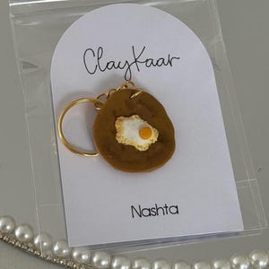 May include: A handmade keychain featuring a fried egg design. The egg is brown with a white and yellow yolk, attached to a gold-colored keyring. The keychain is on a white card with the text "Clay Kaar" and "Nashta".
