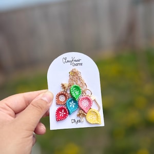 May include: A necklace featuring colorful, teardrop-shaped charms. Each charm is a different color, including red, blue, green, and yellow, with intricate white designs. The card reads "Claykaar Charms" and "Diya".