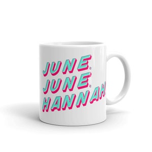 Op de afbeelding: Witte keramische mok met de tekst "JUNE. JUNE. HANNAH" in roze en turquoise letters.