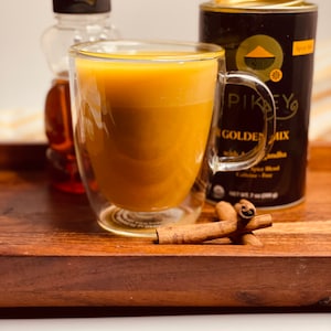 K&ouml;nnte beinhalten: Ein Glas goldene Milch in einem doppelwandigen Glasbecher, eine Flasche Ahornsirup und eine Dose "Pikeys Golden Mix" Gew&uuml;rzmischung auf einem Holztablett mit Zimtstangen.
