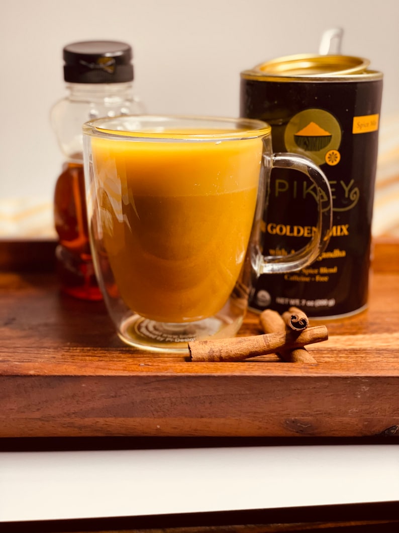 K&ouml;nnte beinhalten: Ein Glas goldene Milch in einem doppelwandigen Glasbecher, eine Flasche Ahornsirup und eine Dose "Pikeys Golden Mix" Gew&uuml;rzmischung auf einem Holztablett mit Zimtstangen.