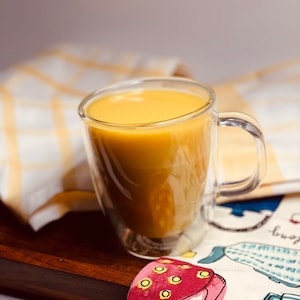 K&ouml;nnte beinhalten: Eine gl&auml;serne Tasse gef&uuml;llt mit einer goldgelben Fl&uuml;ssigkeit, die auf einer Holzoberfl&auml;che mit einem gemusterten Tuch darunter steht.