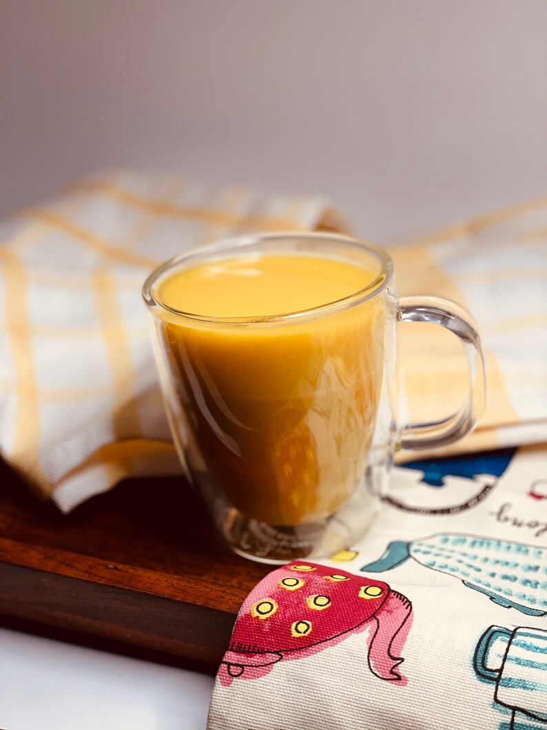 K&ouml;nnte beinhalten: Eine gl&auml;serne Tasse gef&uuml;llt mit einer goldgelben Fl&uuml;ssigkeit, die auf einer Holzoberfl&auml;che mit einem gemusterten Tuch darunter steht.