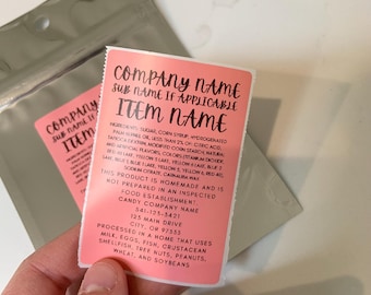 May include: Pink and white label with black text listing ingredients, company name, address, and a statement that the product is homemade and not prepared in an inspected food establishment. The text reads: "COMPANY NAME SUB NAME IF APPLICABLE ITEM NAME INGREDIENTS: SUGAR, CORN SYRUP, HYDROGENATED PALM KERNEL OIL, LESS THAN 25% OF: CITRIC ACID, TAPIOCA DEXTRIN, MODIFIED CORN STARCH, NATURAL AND ARTIFICIAL FLAVORS, COLORS (TITANIUM DIOXIDE, RED 40 LAAF, YELLOW 5 LAKE, YELLOW 6 LAKE, BLUE 2 LAKE, BLUE & BLUE 1 LAKE, YELLOW 5, YELLOW 6, RED 40), SODIUM CITRATE, CARNAUBA WAX THIS PRODUCT IS HOMEMADE AND IS NOT PREPARED IN AN INSPECTED FOOD ESTABLISHMENT. CANDY COMPANY NAME 123 MAIN DRIVE CITY, OR 97555 PROCESSED IN A HOME THAT USES MILK, EGGS, FISH, CRUSTACEAN SHELLFISH, TREE NUTS, PEANUTS, WHEAT, AND SOYBEANS"