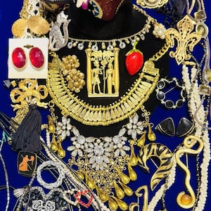 May include: A collection of assorted costume jewelry including gold-tone necklaces, brooches, and earrings. Items feature Egyptian-style motifs, clear crystals, red stones, black enamel, pearls, and a small figurine. Some pieces are on display cards.
