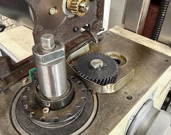 May include: Close-up of industrial machinery components. The image shows a complex arrangement of gears, including a small brass gear and a larger black gear, mounted on a metal surface. The machinery appears to be well-used, with visible wear and tear.