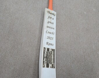 May include: A white wooden baseball bat with an orange handle. The bat has a black and white photo of a team on it. The text on the bat reads "Thanks for a great season Coach! 2025 Rams".