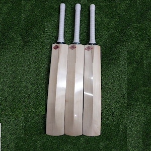 Puede incluir: Tres bates de cricket de madera con empuñaduras blancas y un logotipo rojo. Los bates están dispuestos uno al lado del otro sobre una superficie de césped artificial verde.