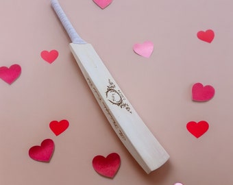 May include: A wooden cricket bat with a white grip and a personalized inscription. The bat is surrounded by pink and red hearts on a light pink background.