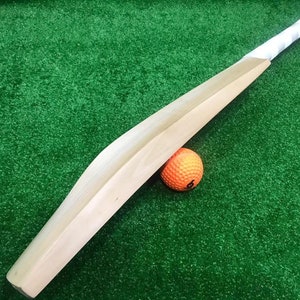 May include: A wooden cricket bat with a white grip, lying on a green artificial grass surface with an orange cricket ball beside it.