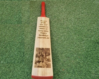May include: A wooden cricket bat with a red handle and a personalized message engraved on the front. The message reads "40 years of life, love and adventure. Here's to many more. Happy Birthday! 2023-03-22". The bat also features a photo of a group of people celebrating a birthday.