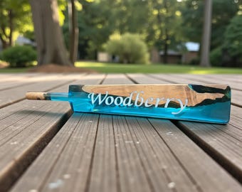 May include: A handcrafted bottle stopper with a wooden and blue resin design. The stopper features the word "Woodberry" in white script. The wooden portion is light brown, and the resin is a vibrant blue. The stopper has a cork end.