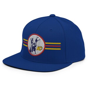 May include: Royal blue flat-brim baseball cap with an embroidered patch. The patch features a white circle with a blue and white image of a horse and rider, with the letters "KC" in gold. Two horizontal stripes in red and gold are also embroidered.