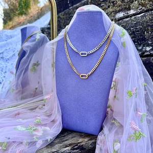 May include: Two chain necklaces displayed on a purple bust. One necklace is gold-toned, the other is silver-toned. Both feature a curb chain design with an oval clasp. The bust is draped with sheer fabric with floral embroidery.