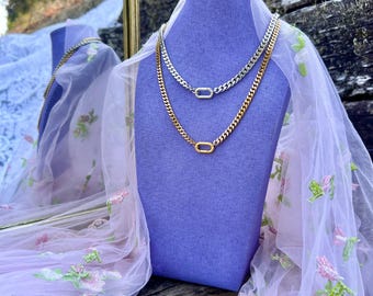 Puede incluir: Dos collares de cadena exhibidos en un busto morado. Uno es dorado, el otro plateado. Ambos presentan un diseño de cadena de eslabones con un cierre ovalado. El busto está cubierto con tela transparente con bordados florales.