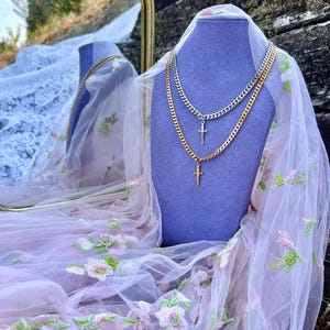 May include: Two necklaces with cross pendants displayed on a purple bust. One necklace is gold-toned, the other silver-toned. The necklaces are on a bust covered with a sheer, floral-embroidered fabric. A gold-framed mirror is in the background.
