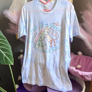 K&ouml;nnte beinhalten: Ein hellblaues Batik-T-Shirt mit einer Grafik von zwei Cartoon-Pok&eacute;mon-Charakteren. Der Text "Only Gay People Can Read This" ist in einem Regenbogenbogen um die Figuren gedruckt.