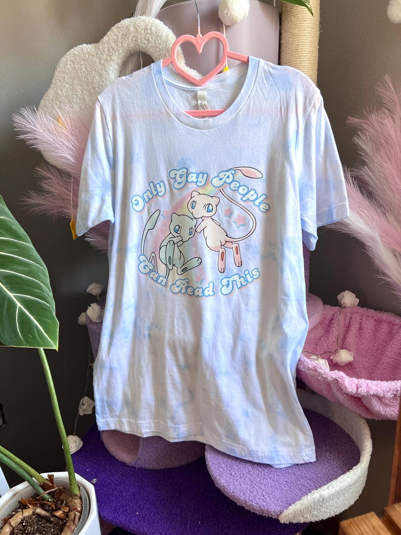 K&ouml;nnte beinhalten: Ein hellblaues Batik-T-Shirt mit einer Grafik von zwei Cartoon-Pok&eacute;mon-Charakteren. Der Text "Only Gay People Can Read This" ist in einem Regenbogenbogen um die Figuren gedruckt.