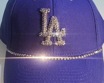 May include: A royal blue baseball cap with a white brim and a silver rhinestone LA logo. The cap is decorated with a row of rhinestones around the brim.