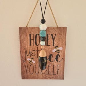 Puede incluir: Un letrero de madera con el texto "Honey just bee yourself" e ilustraciones de abejas. Un cord&oacute;n negro con cuentas de colores y un difusor negro cuelgan de la parte superior. Las cuentas son negras, blancas, turquesas y grises.