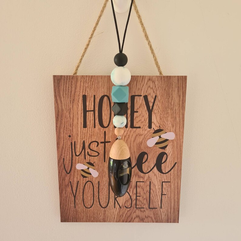 Puede incluir: Un letrero de madera con el texto "Honey just bee yourself" e ilustraciones de abejas. Un cord&oacute;n negro con cuentas de colores y un difusor negro cuelgan de la parte superior. Las cuentas son negras, blancas, turquesas y grises.