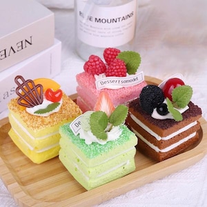 Puede incluir: Cuatro bizcochos cuadrados en miniatura sobre una bandeja de madera. Los bizcochos son amarillos, rosas, verdes y marrones, cada uno cubierto con diferentes frutas y elementos decorativos. El bizcocho rosa tiene el texto "Dessert somodel".