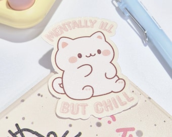 Könnte beinhalten: Ein hellrosa Aufkleber mit einer Cartoon-Katze und dem Text "MENTALLY ILL BUT CHILL". Der Aufkleber befindet sich auf einer hellen Oberfläche mit einem Stift und einem Taschenrechner im Hintergrund.
