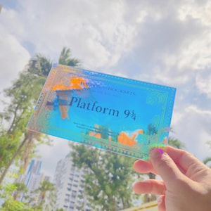 Peut inclure: Un billet holographique avec le texte "London to Hogwarts" et "Platform 9 3/4" dans un design bleu et or. Le billet a un effet chatoyant et irisé, avec un fond de ciel nuageux et de palmiers.