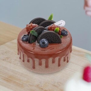 Pode incluir: Um bolo redondo de chocolate com uma cobertura castanha escura escorrendo pelas laterais. O bolo é coberto com biscoitos Oreo, mirtilos e frutos silvestres vermelhos. Uma pequena etiqueta branca com a palavra "odel" é inserida no bolo.