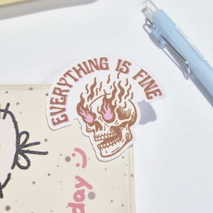 Może przedstawiać: Naklejka z brązową czaszką z różowymi płomieniami i napisem "EVERYTHING IS FINE" w zakrzywionym banerze. W tle widoczny jest jasnoniebieski długopis.
