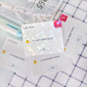 May include: Holographic stickers with a computer error theme. Each sticker has a white background with text such as "Life Error" and "no motivation found." The stickers also feature an "OK" button and an exclamation mark.