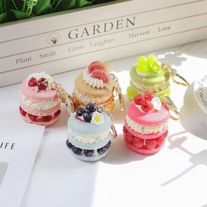 May include: A collection of colorful macaron keychains. Each keychain features a miniature, layered macaron design with various toppings like berries and cream. The keychains are in shades of pink, blue, green, and tan, with gold-colored key rings.
