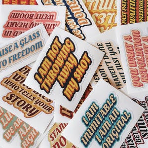 May include: A collection of white stickers with various phrases in retro fonts. The stickers feature phrases like "I am not throwing away my shot" and "Who tells your story". The color palette includes orange, yellow, blue, and red.
