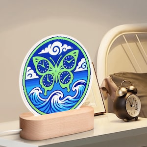 Puede incluir: Una luz de noche decorativa con un diseño de mariposa. Las alas de la mariposa presentan esferas de reloj, sobre un fondo azul y blanco con olas y nubes. La luz se apoya en una base de madera.