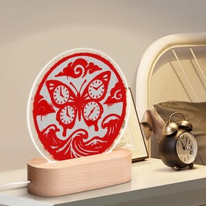 Puede incluir: Luz de noche decorativa con un diseño de mariposa roja que incorpora esferas de reloj en un panel redondo e iluminado. Se apoya en una base de madera y tiene un cable de alimentación blanco. El diseño incluye nubes y olas.