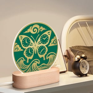 Puede incluir: Luz de noche decorativa con un diseño de mariposa verde y dorada y esferas de reloj, colocada sobre una base de madera. El diseño de la mariposa está sobre un fondo verde con detalles dorados. La luz se alimenta con un cable blanco.