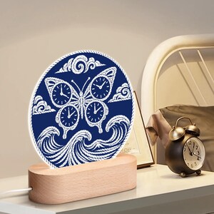 Puede incluir: Luz de noche decorativa con un diseño de mariposa azul y blanco, que incorpora esferas de reloj, sobre una base de madera. El diseño de la mariposa está rodeado de nubes y olas. La luz se alimenta con un cable blanco.