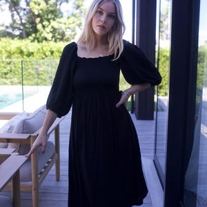 Puede incluir: Una mujer lleva un vestido midi negro con escote cuadrado y mangas abullonadas. El vestido tiene un corpi&ntilde;o fruncido y una falda fluida. Est&aacute; de pie en una terraza de madera con una piscina en el fondo.