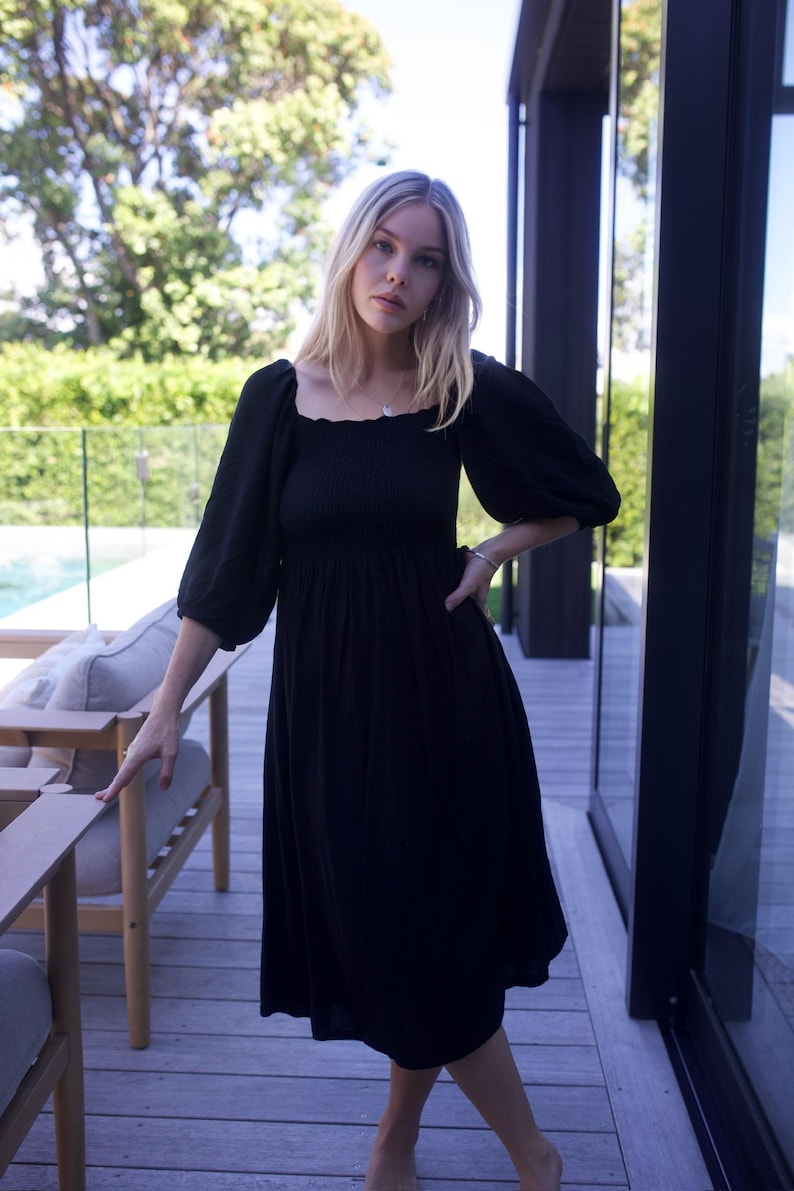 Puede incluir: Una mujer lleva un vestido midi negro con escote cuadrado y mangas abullonadas. El vestido tiene un corpi&ntilde;o fruncido y una falda fluida. Est&aacute; de pie en una terraza de madera con una piscina en el fondo.