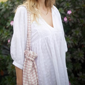 May include: A woman wearing a white, embroidered, loose-fitting blouse with a pink and white gingham tote bag.