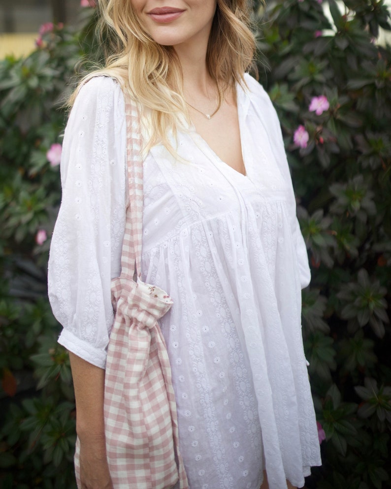 May include: A woman wearing a white, embroidered, loose-fitting blouse with a pink and white gingham tote bag.