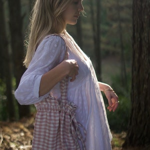 May include: A woman in a white dress with a pink and white gingham bag slung over her shoulder. She is standing in a forest setting.