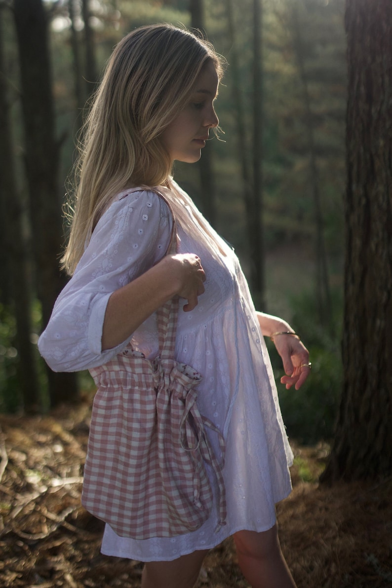 May include: A woman in a white dress with a pink and white gingham bag slung over her shoulder. She is standing in a forest setting.