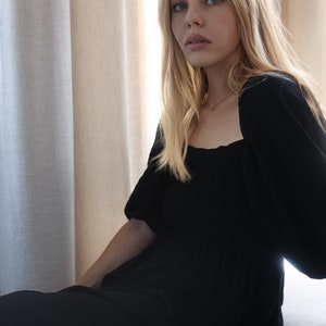 Puede incluir: Una mujer con el pelo largo y rubio lleva un vestido negro de cuello cuadrado con mangas abullonadas. Est&aacute; sentada frente a una ventana con cortinas blancas.